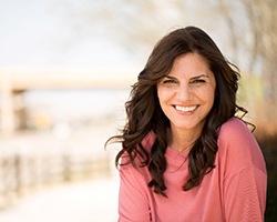 A middle-aged woman wearing a pink blouse with long, dark hair smiles after receiving her dental crowns in Weyauwega