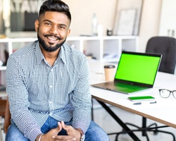 A young man wearing a plaid button-down shirt and sitting at a computer smiling after undergoing multiple tooth extractions in Weyauwega and receiving platelet rich fibrin