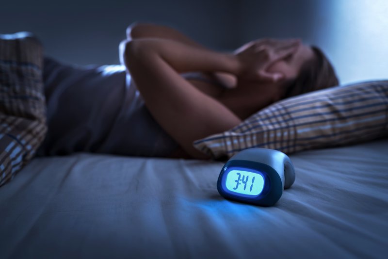 an individual lying in bed covering their face while the alarm clock reads 3:41 a.m.